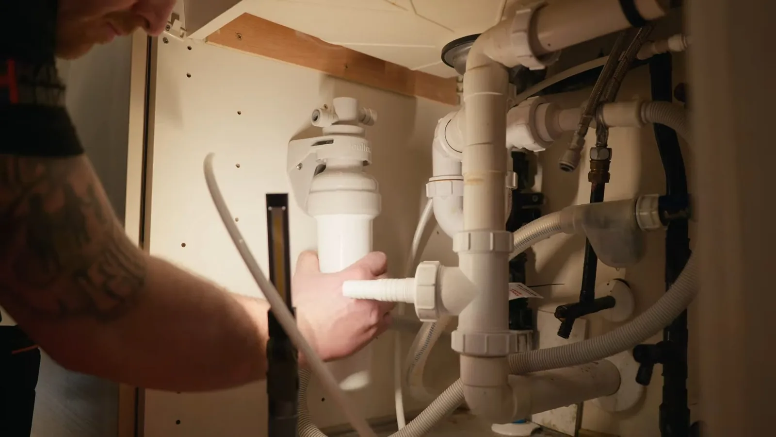 Hand fitting white tubing to a small housing among white drain pipes in an open under-sink cabinet