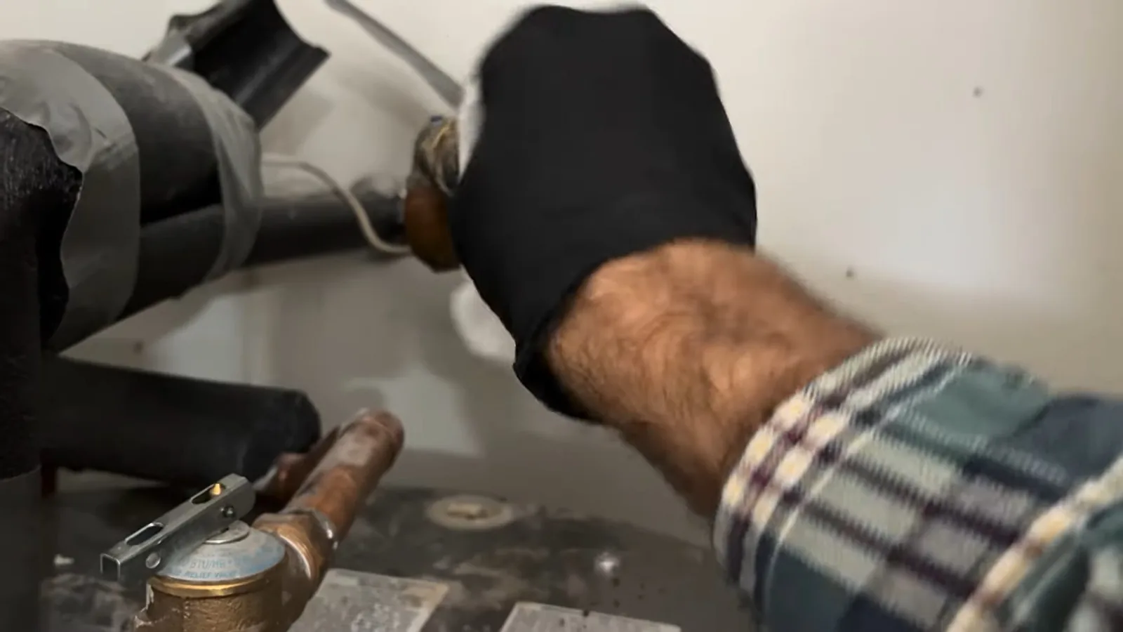 Gloved hand in a plaid sleeve working at the inlet hose and brass valve on top of a water heater
