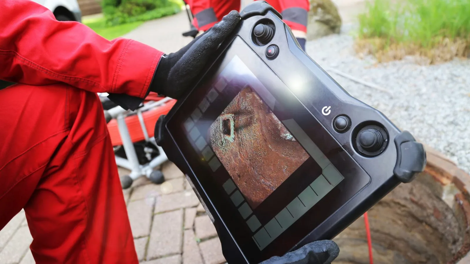 Technician in red coveralls holding a handheld camera monitor that shows the inside of a pipe, beside an open cleanout in a paved yard