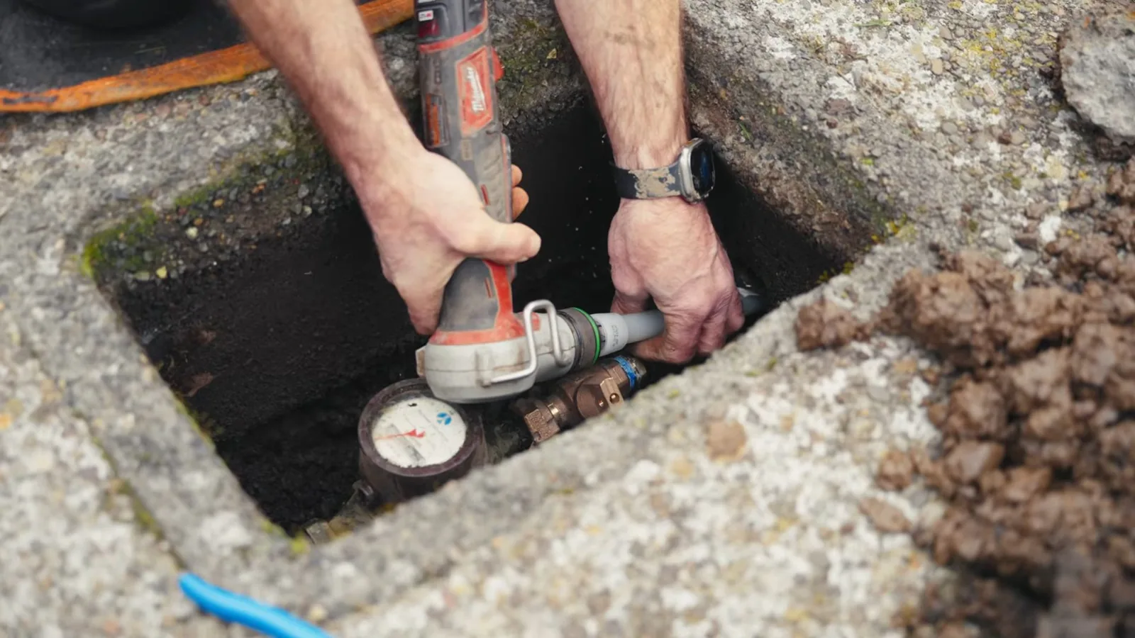A technician's hands working with a tool inside an in-ground meter box, with the water meter visible at the bottom