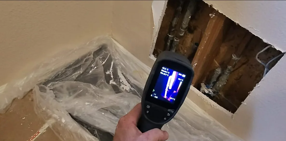 Hand holding a thermal imaging camera in front of an opened wall section that shows pipes, with the screen showing a hot and cold reading