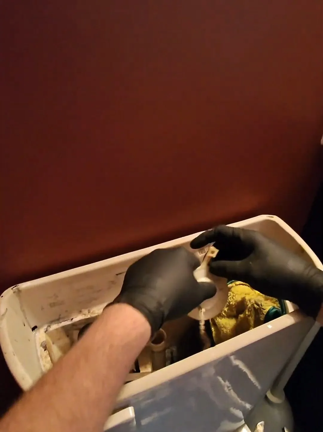 Gloved hand holding a worn part inside an open toilet tank against a dark red wall