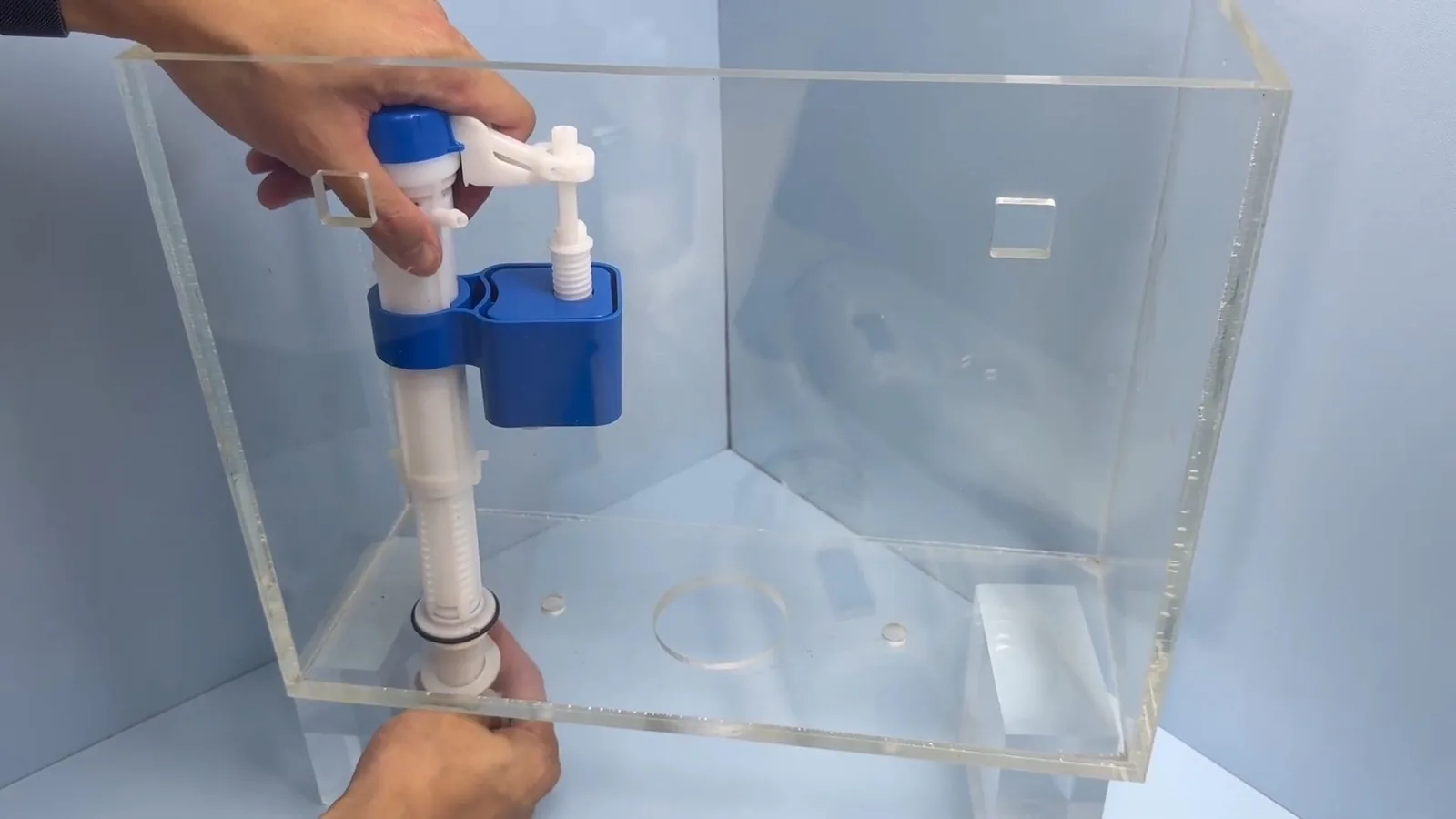 Hands fitting a blue and white fill valve and flush kit into a clear plastic toilet tank model