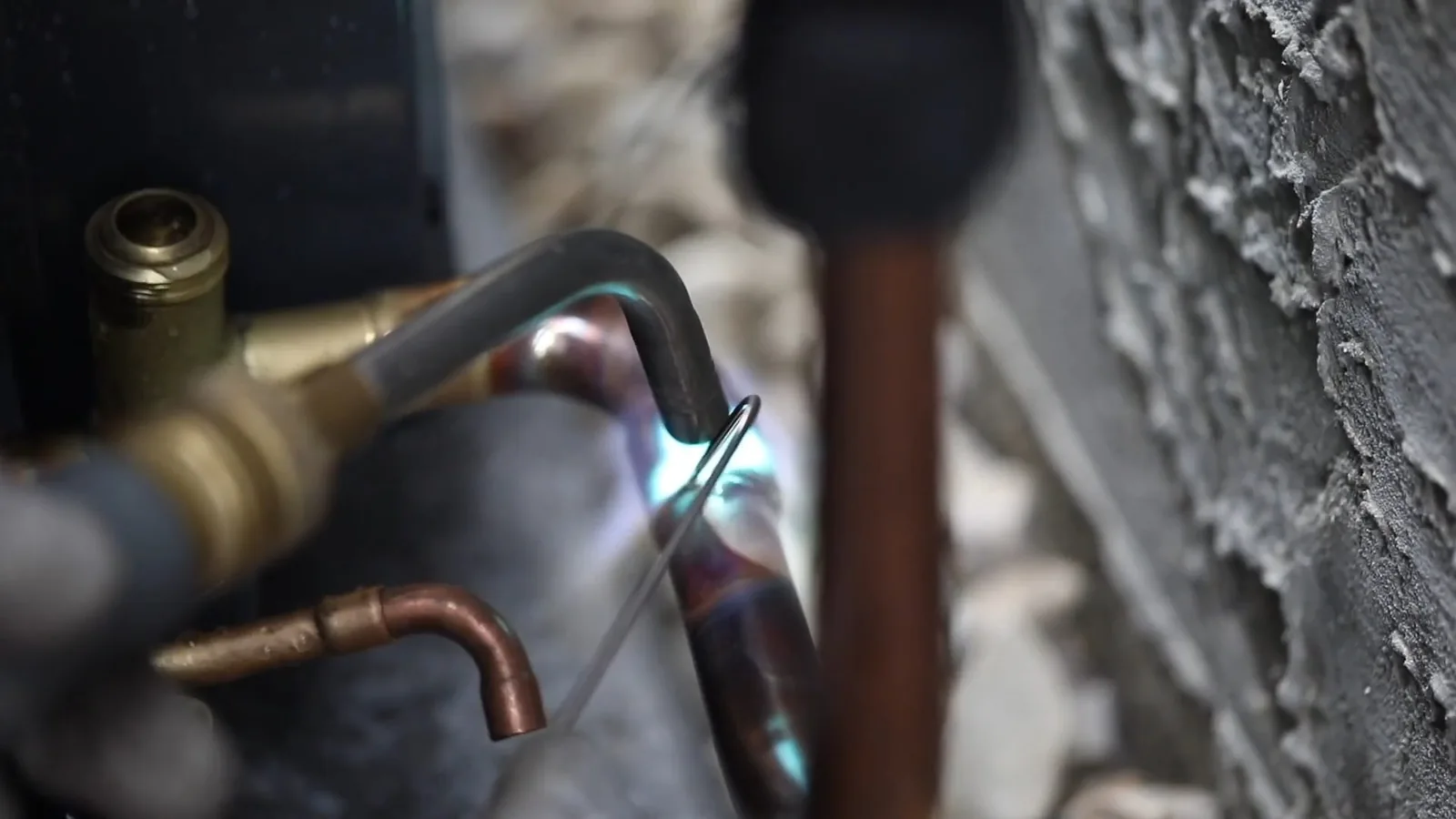 Blue flame from a torch heating a copper fitting at a water heater connection, with a brass valve in the foreground and a stucco wall behind