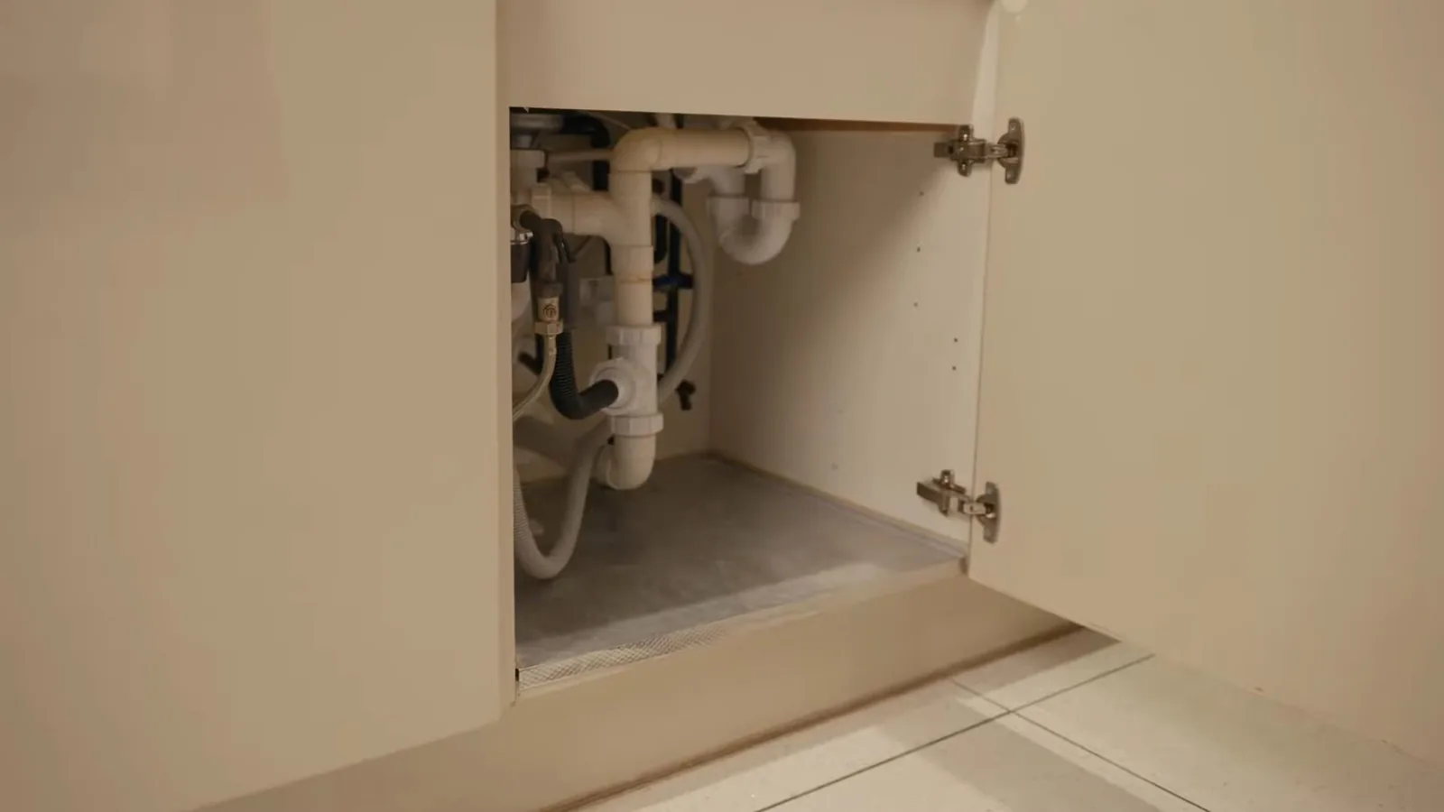 Open cabinet under a sink showing white drain pipes, a trap and supply tubing against a cream interior