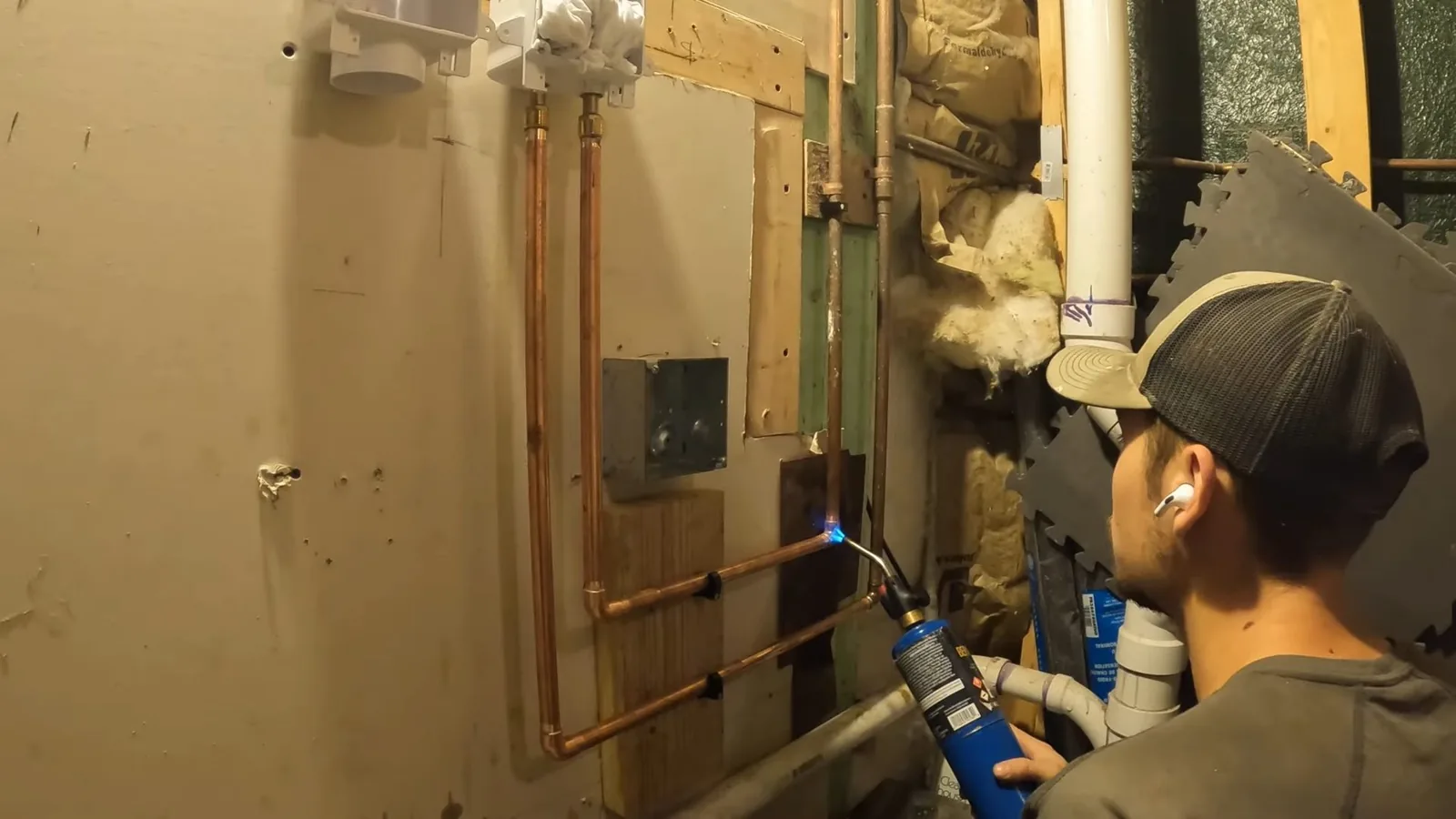 Person holding a torch at copper pipes beside a laundry box and white drain pipe in an open basement wall