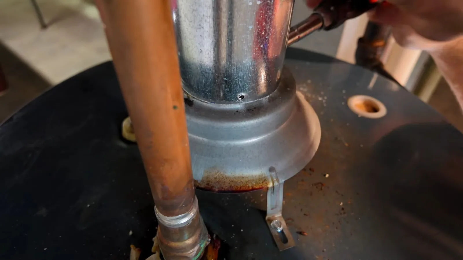 Galvanized vent pipe and collar fitted above a black-topped water heater, with a copper pipe on the left and a hand reaching in at the top right