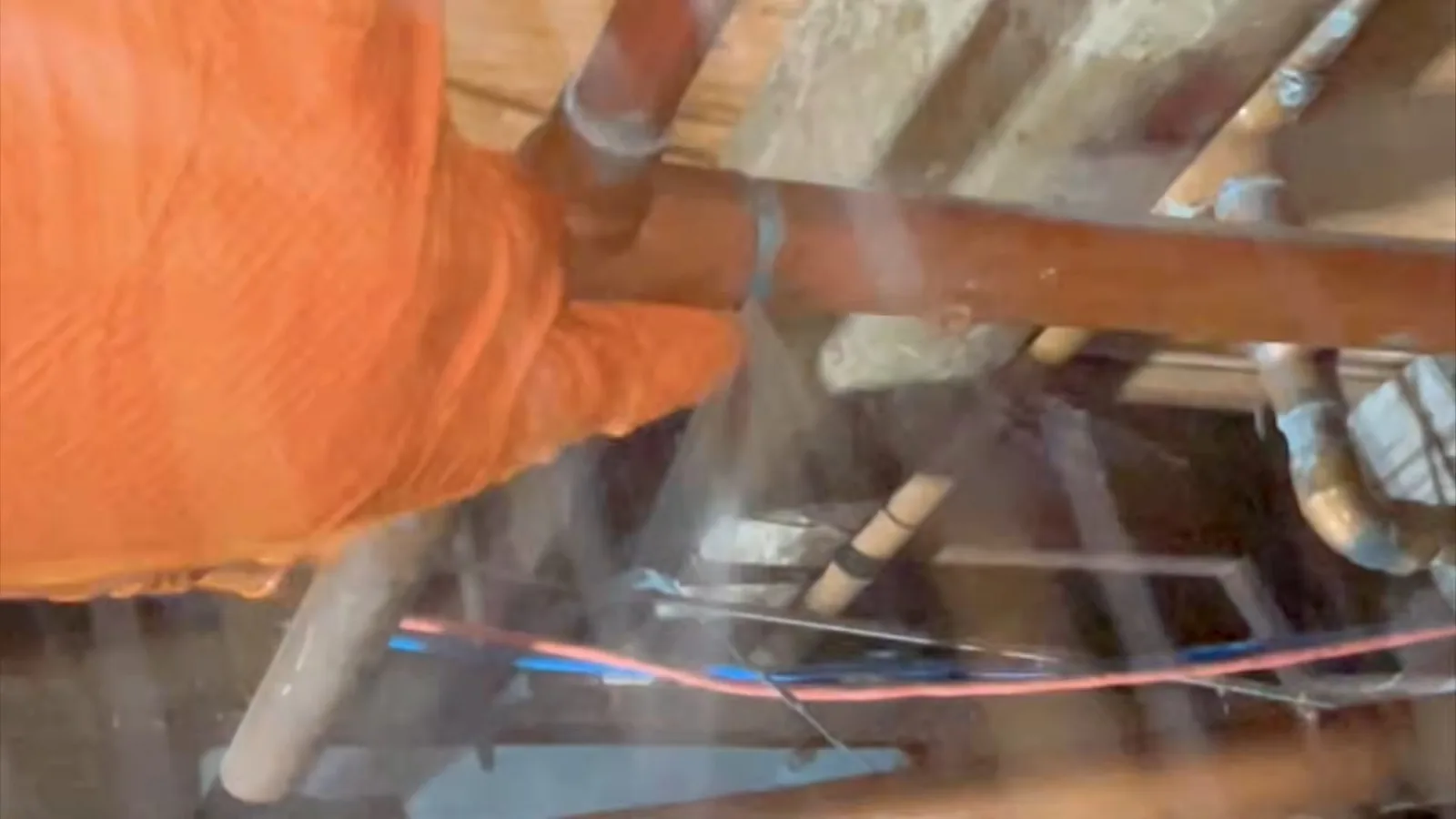 Orange-gloved hand beside a copper pipe under a floor where water is spraying from the break, with wood joists and wiring behind