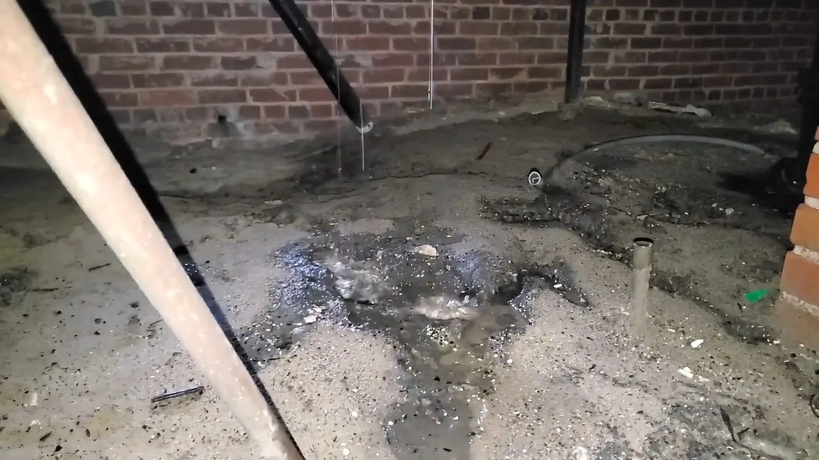 Dark, wet soil with standing water on the floor of a crawlspace, beneath a leaking pipe, with a brick wall and a wooden beam behind
