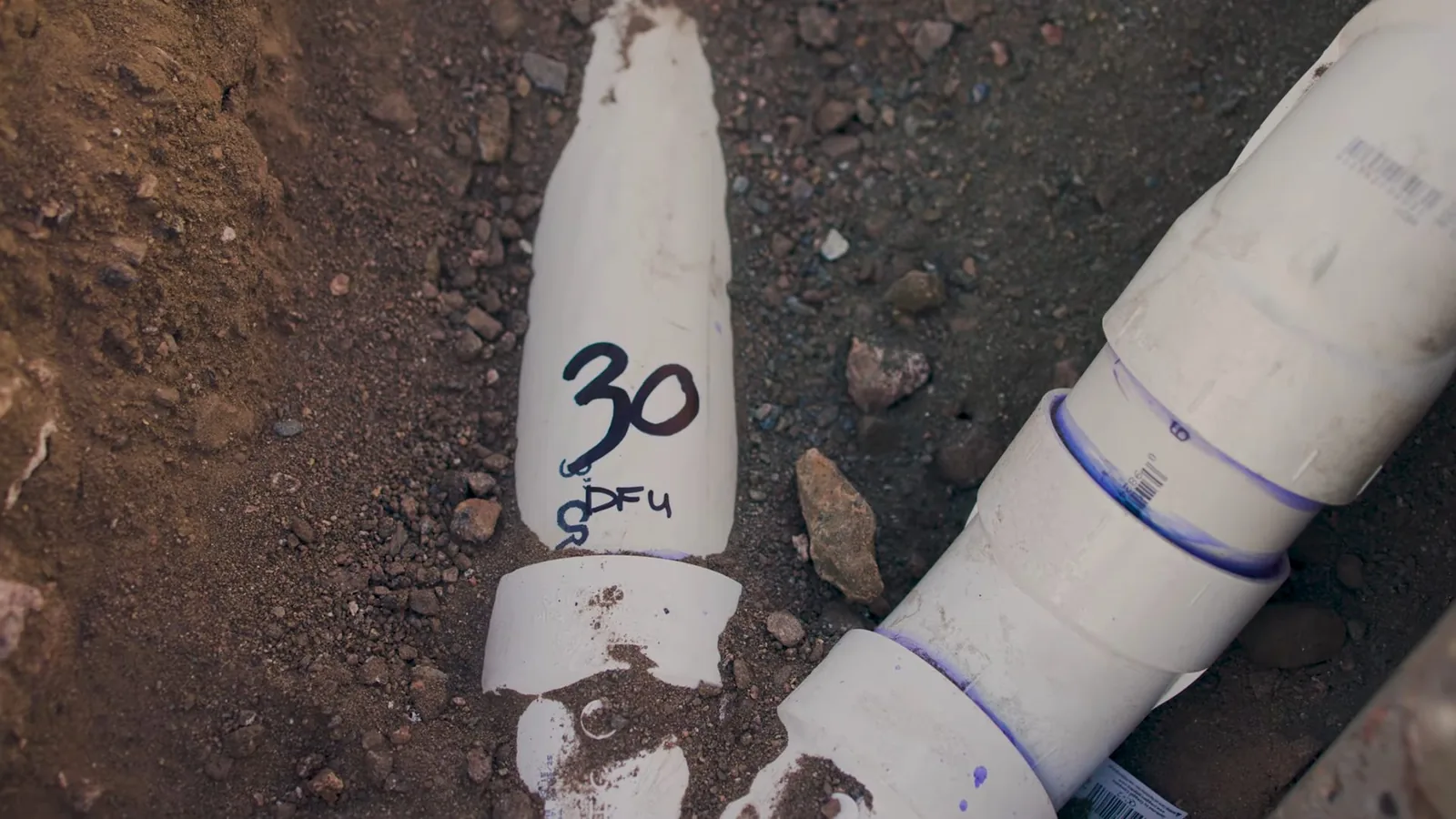 White PVC drain pipe sections with blue primer joints and a hand-written number 30 on one, in bare soil