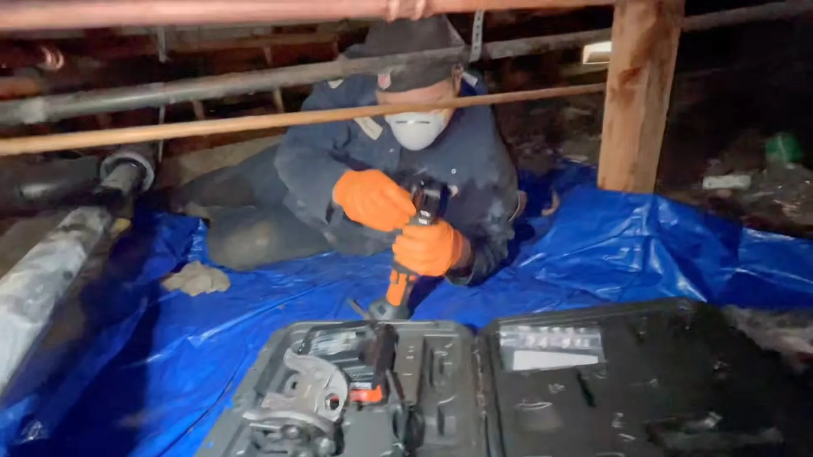 Worker in a dust mask and orange gloves kneeling on a blue tarp in a crawlspace, using a power tool on a pipe above an open tool case