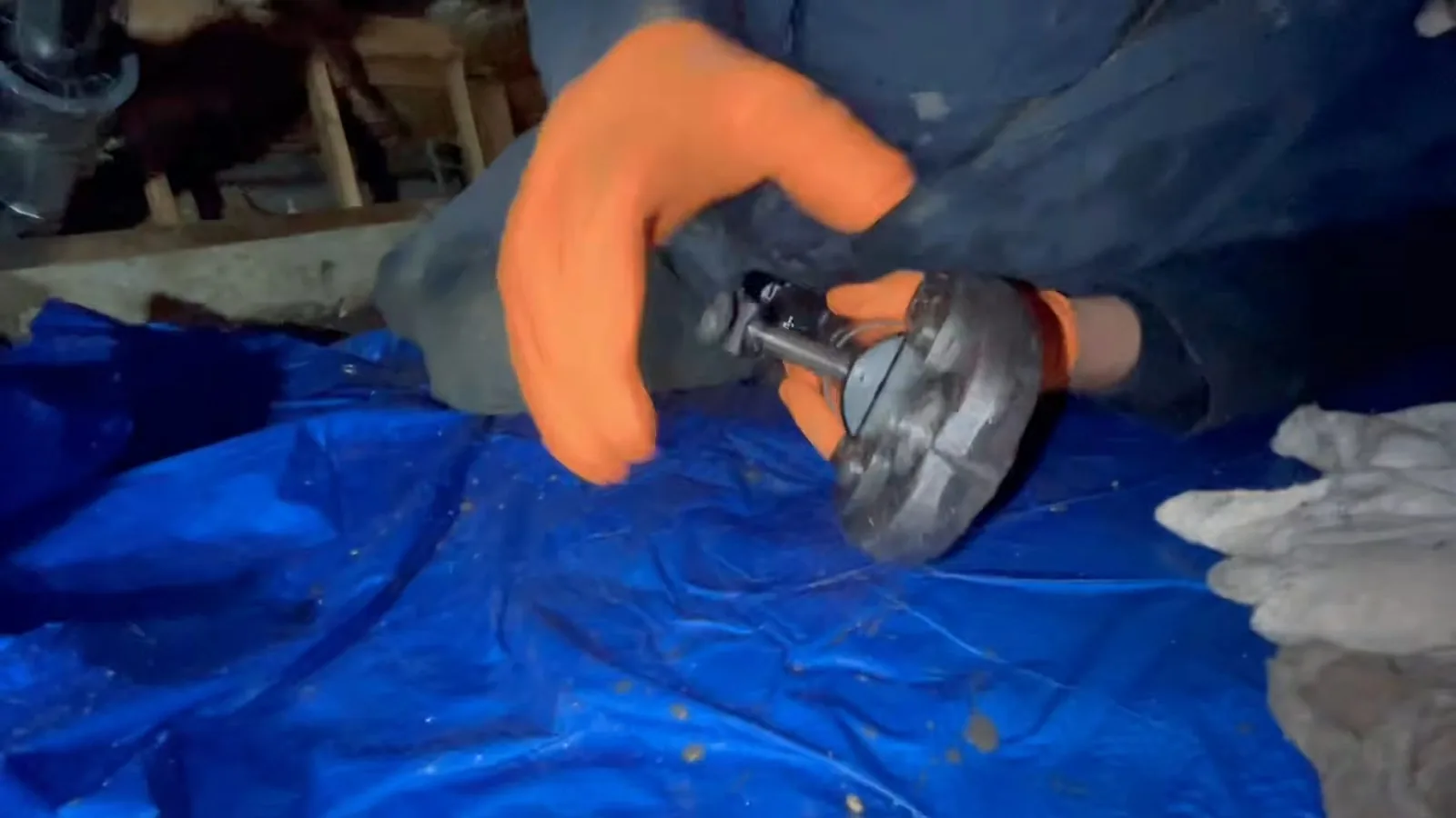 Worker in orange gloves holding a pipe cutter above a blue tarp in a crawlspace